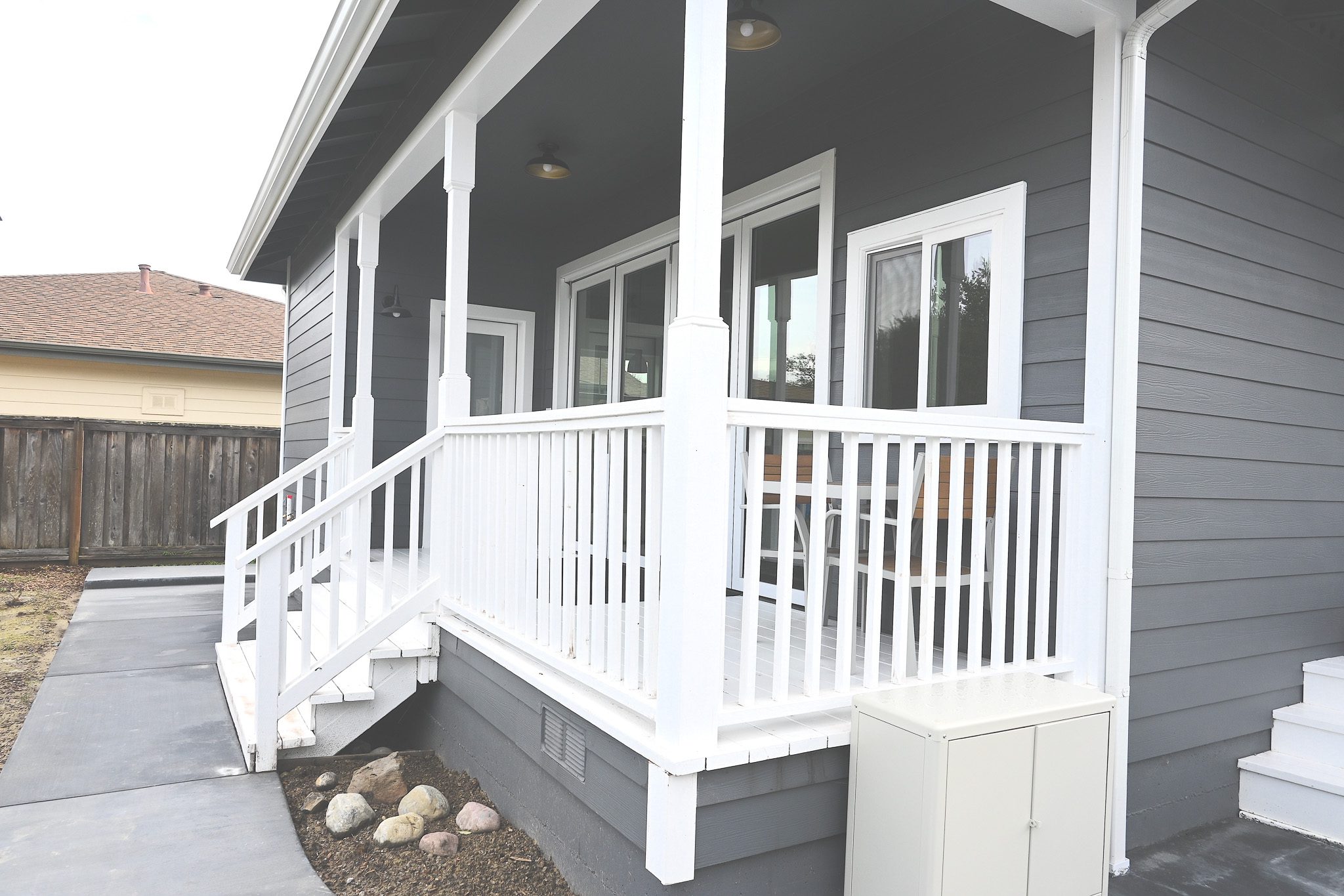 A gray house with white trim features a large white wooden porch with railing and steps. A small shrub garden is visible beside the porch. A wooden fence and an adjacent house are in the background.