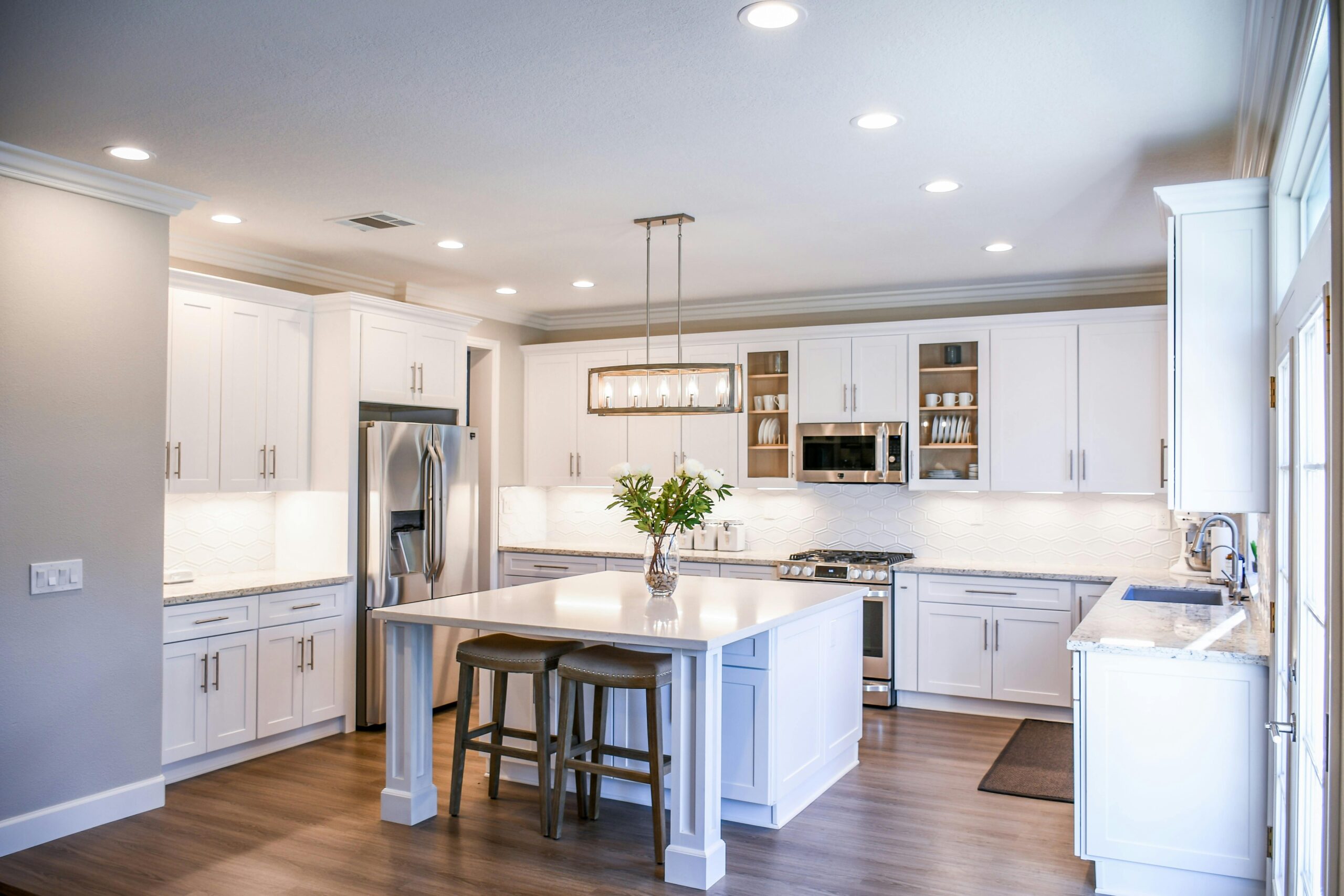 Modern kitchen with white cabinets, stainless steel appliances, an island with two stools, wood flooring, and pendant lighting above the island.